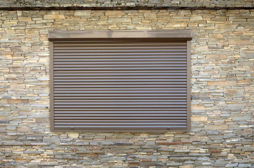 Persiana enrollable exterior, ventana con persianas enrollables de metal marrón. Decoración de pared con piedra aplanada artificial.