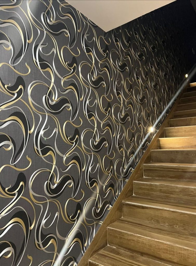 Papel tapiz color gris, con detalles de lineas curveadas de color negro, platado y dorado, colocado en pasillo de escaleras.