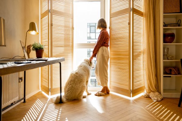 Persianas Monterrey; una señora junto a su mascota viendo el exterior de su habitación con unas persianas que resaltan en la decoración de su hogar.